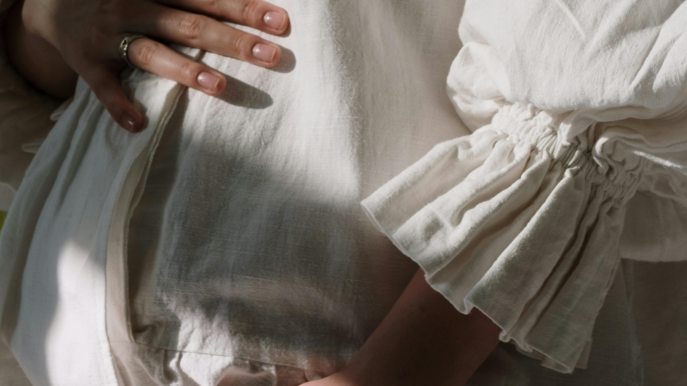 Mains posées sur un ventre de femme enceinte vêtue de lin blanc – illustration de la douceur et du bien-être pendant la grossesse, Oca Rina.