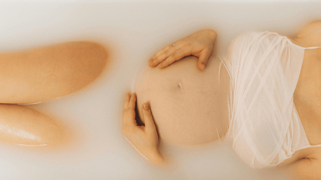 Femme enceinte allongée dans un bain de lait, mains posées sur son ventre arrondi – moment de détente et de connexion à la maternité, Oca Rina.